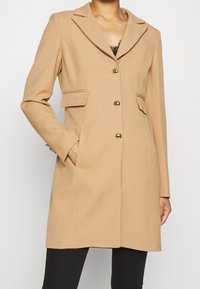 Cappotto beige con taglio su misura, colletto a revers, due tasche frontali e dettagli con bottoni dorati, realizzato in un tessuto morbido.