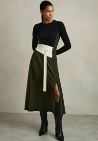 Lange jurk met een zwart geribbelde top en een olijfgroene rok, voorzien van een witte riem met een gesp en een hoge zijsplit. Zwarte laarzen tot de knie.