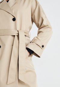 Gabardina beige con un diseño de doble botonadura, solapas anchas y cinturón atado en la cintura. Presenta botones negros y una textura suave.