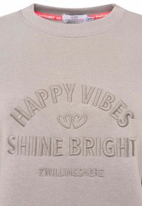 Helles beige Sweatshirt mit geprägtem Text: "HAPPY VIBES" und "SHINE BRIGHT." Rundhalsausschnitt mit geripptem Strickmuster. Markenetikett sichtbar.