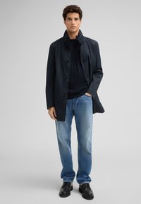 Marineblaue, leichte, knielange Jacke mit Knopfverschluss, kombiniert mit hellblauen Jeans und schwarzen Schnürschuhen. Glatte Textur und lässige Passform.