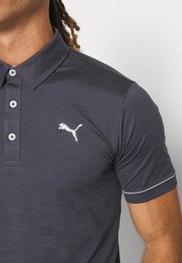 Tmavě šedé polo tričko s texturovaným povrchem, tříknoflíkovým zapínáním a bílými detaily prošívání. Na hrudi se nachází bílé logo Puma.