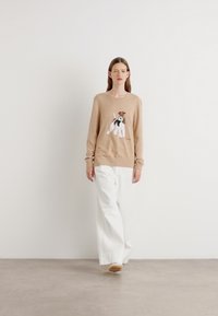 Pulover tricotat bej cu un grafic de câine pe față, asortat cu pantaloni albi largi și încălțăminte de culoare deschisă. Design simplu și casual.