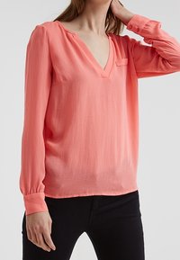 Blusa a maniche lunghe color corallo con scollo a V e tasca sul petto. Tessuto leggero con una texture liscia e polsini arricciati. Pantaloni neri visibili.