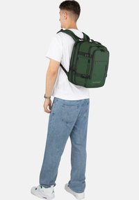 Grüner Rucksack aus strapazierfähigem Stoff, mit mehreren Fächern, schwarzen Akzenten und verstellbaren Trägern, auf dem Rücken einer Person getragen.
