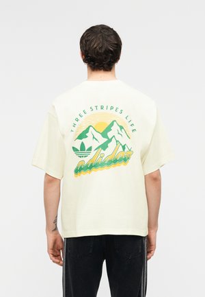 Mężczyzna noszący kremowy t-shirt z zielono-żółtą grafiką gór oraz napisami "Three Stripes Life" i "adidas" na plecach.