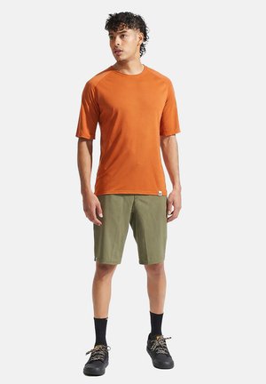 Junger Mann steht, trägt orangefarbenes kurzärmeliges Hemd, olivgrüne Shorts, schwarze Socken und schwarze Turnschuhe, schaut leicht nach links.