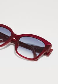 Ochelari de soare roșii cu lentile gradient, cadrul din plastic lucios și branding minim pe brațe. Design unghiular cu o lățime notabilă.