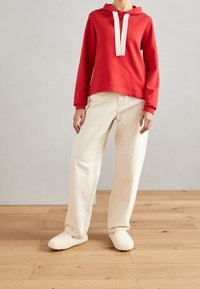 Sudadera roja con cordones blancos, combinada con pantalones sueltos de color crema y suaves zapatillas de color blanco roto. Presenta un diseño casual y un ajuste relajado.