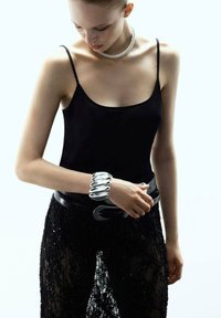 Mujer con top negro sin mangas y pantalones de encaje, llevando un collar de plata grueso y múltiples pulseras de plata, ajustándose el cinturón.