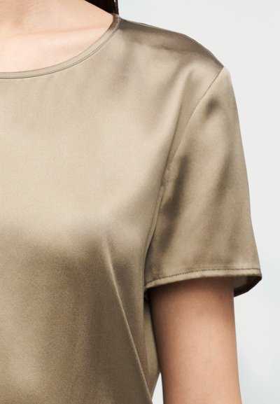 Κοντομάνικη μπλούζα από απαλό, ελιά πράσινο ύφασμα με λεία υφή και στρογγυλή λαιμόκοψη. Απλός σχεδιασμός, χωρίς ορατά σχέδια ή μεταλλικά στοιχεία.
