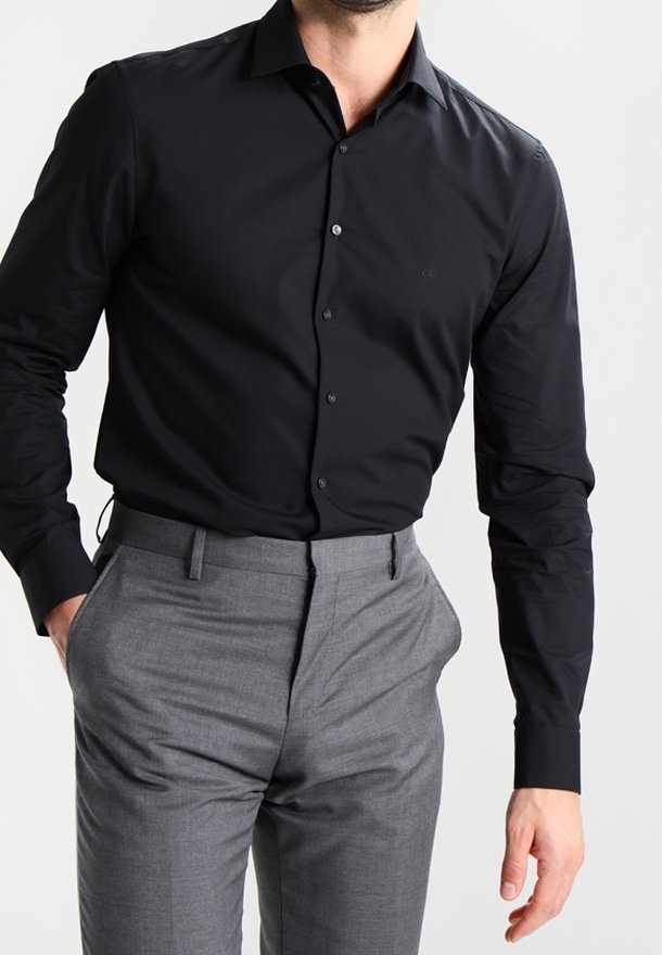 Man in een goed passende zwarte overhemd met knopen, in de hoge taille grijze broek gestopt, met zijn handen dicht bij de zakken.