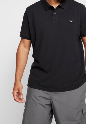 Mand iført en sort kortærmet polo med et lille GANT-logo, kombineret med grå cargobukser mod en ensfarvet baggrund.