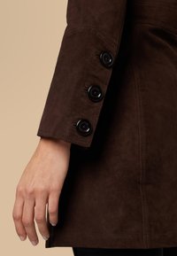 Brown suede coat sleeve with black buttons, featuring a smooth texture, tailored fit, and visible stitching detail at cuff and seam.