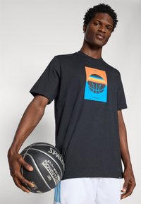 T-shirt noir à manches courtes avec un graphique de basket-ball coloré sur le devant, porté par une personne tenant un ballon de basket noir.