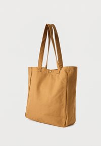 Καφέ υφασμάτινη τσάντα ώμου με δύο μακριές λουριά και μικρό μεταλλικό κουμπί κλεισίματος, εμφανίζεται πάνω σε απλό φωτεινό φόντο.