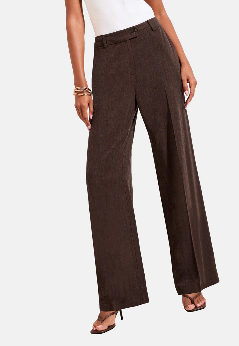 Woman wearing high-waist wide-leg brown pants, white top, bracelets, and brown heeled sandals standing against a plain background.