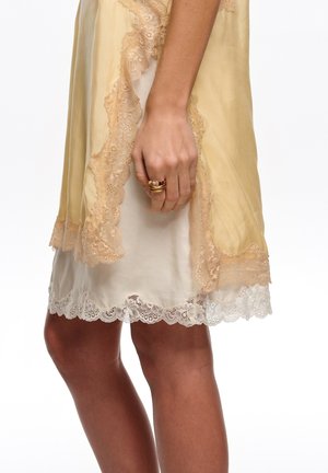 Hand with multiple gold rings resting against cream slip dress with white lace hem and sheer gold lace-trimmed robe over it.