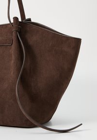 Ruskea nubukkikangas käsilaukku, jossa on pehmeä pinta, pitkänomainen muoto ja ohut nahkaremmi. Laukussa on minimalismi ja siinä ei ole näkyvää metalliosaa.