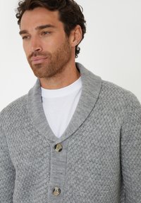 Mann trägt eine graue strukturierte Strickjacke mit Schalkragen und großen Knöpfen über einem weißen Rundhalsausschnitt-T-Shirt vor weißem Hintergrund.