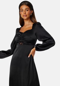 Černé saténové šaty se čtvercovým výstřihem, nařasením v oblasti prsou, jemnými krajkovými detaily a nafouklými dlouhými rukávy. Hladká textura, minimalistický design.