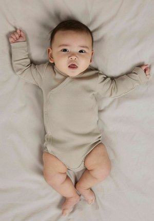 Bodysuit pour bébé beige clair à manches longues en tissu côtelé doux avec des fermetures à pression sur le côté, posé sur une surface de couleur claire.