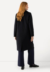 Dlouhý námornický kabát se rovný střih, bočními rozparky a límečkovým výstřihem. Nosený přes široké námornické kalhoty a černé mokasíny.