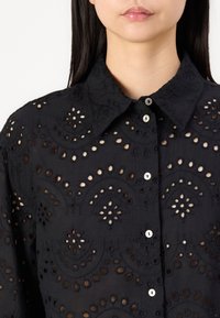 Black embroidered blouse featuring cut-out patterns, a classic collar, and button closure, with a textured fabric and intricate detailing.