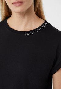 Schwarzes Baumwoll-T-Shirt mit runder Ausschnitt. Mit gesticktem weißen Text "NUR GUTE VIBEN" in der Nähe des Kragens. Kurze Ärmel.
