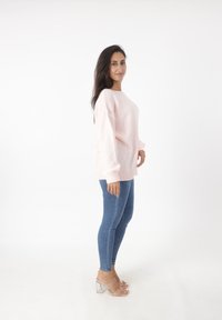 Lichtroze gebreide trui met een losse pasvorm, geribbelde manchetten, gedragen over strakkere blauwe jeans en doorzichtige hakken. Losse achtergrond.