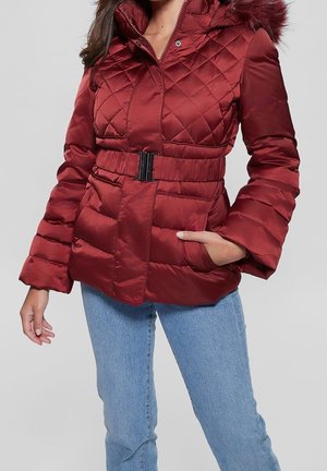 Femme portant une veste d'hiver matelassée rouge avec ceinture et capuche bordée de fourrure, associée à un jean bleu clair, mains détendues dans les poches.