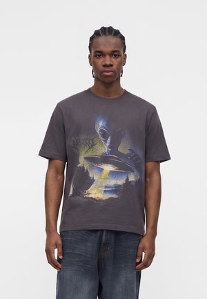 Νέος άνδρας με πλεξούδες φοράει σκούρο γκρι μπλουζάκι με μπλε εξωγήινο και UFO που εκπέμπει φως πάνω από σκηνή δάσους.