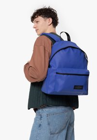 Blå nylonryggsäck med en framficka och svart dragkedja, bärs av en person i en vadderad väst, och visar en rundad form och slät textur.