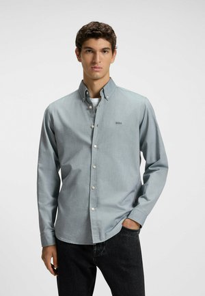 Camisa de botones azul claro con cuello, tela texturizada, bolsillo frontal y un pequeño logo "BOSS" en el lado izquierdo. Combinada con jeans oscuros.