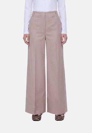 Pantaloni beige a gamba larga con sottili righe bianche verticali e polsini rigati orizzontali, indossati con un top bianco a maniche lunghe e scarpe rigate.