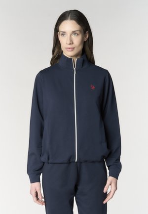 Donna che indossa una giacca blu navy con zip, collo alto e piccolo logo rosso ricamato sul petto sinistro, in piedi davanti a uno sfondo uniforme.