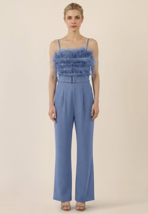 Femme en combinaison bleu sans manches avec corsage plumé et ceinture, debout devant un fond beige uni.