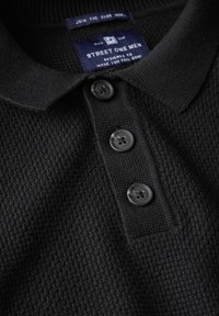 Schwarzes Poloshirt aus strukturiertem, gewebtem Stoff, mit einem flachen Kragen und drei dekorativen Knöpfen. Das Etikett trägt die Aufschrift "Street One Men."