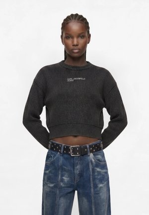 Kvinde med flettet hår iført en sort cropped sweater med teksten "Karl Lagerfeld Jeans" og højtaljede blå jeans med et sort nittebælte.