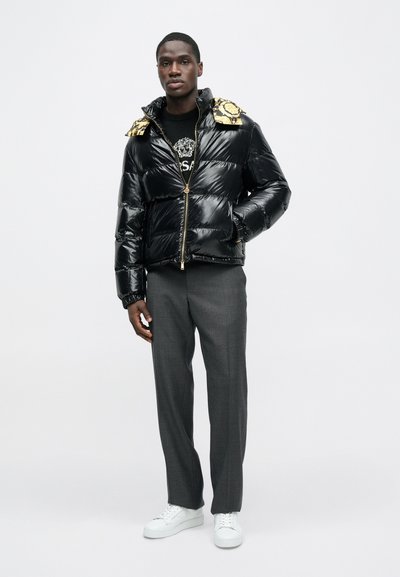 Sort pufferjakke med skinnende tekstur, guld accenter på skuldrene, lynlåslukning, båret over en sort grafisk sweatshirt, kombineret med grå bukser og hvide sneakers.