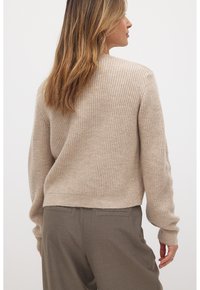 Maglione a coste beige con maniche lunghe e orlo corto, caratterizzato da una finitura testurizzata e un design minimalista. Abbinato a pantaloni grigi.
