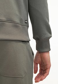 Felpa grigia con polsini a coste, dotata di etichetta. Primo piano della texture del tessuto e della vestibilità attorno al polso e all'area della tasca.
