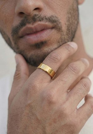 Nahaufnahme von dem unteren Teil eines Mannesgesichts mit kurzem Gesichtshaar, der mit der Hand, an der ein breiter goldener Ring am Mittelfinger sitzt, sein Kinn berührt.