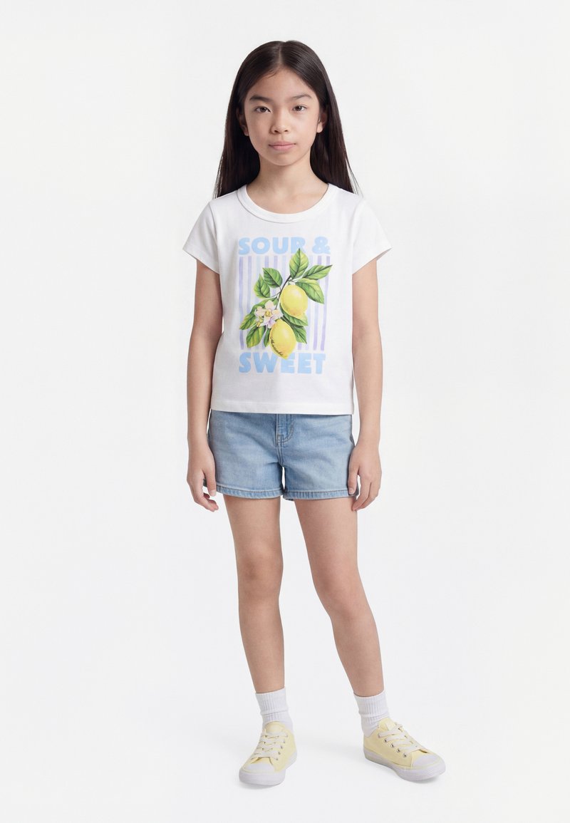 Νεαρή κοπέλα που στέκεται φορώντας λευκή μπλούζα με λεμόνια και το κείμενο «Όξινο & Γλυκό», τζιν σορτς, λευκές κάλτσες και ανοιχτό κίτρινα αθλητικά παπούτσια.