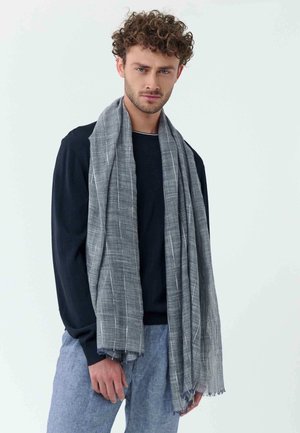 Young man with curly hair wearing black long-sleeve shirt, light blue pants, and a long gray striped scarf against a plain background.