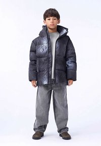 Must paks sulejope tekstuurse pinnaga ja kapuutsiga, kantud heledat halli särgi ning tumedate teksapükstega, kombineeritud tumedate libisemiskingadega.