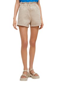 Kobieta ubrana w beżowe szorty o wysokim stanie z podwiniętym dołem oraz masywne beżowe sandały, stojąca na białym tle.