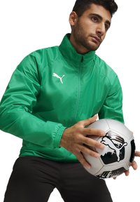 Veste synthétique verte avec col haut, arborant un logo Puma blanc. Le modèle tient un ballon de football noir et argent avec un design graphique.