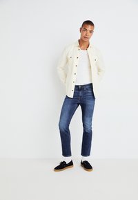 Weißes Hemd mit schwarzen Knöpfen, über einem weißen Tanktop getragen; dunkelblaue Jeans; schwarze Schuhe mit Gummisohlen; einfarbiger weißer Hintergrund.