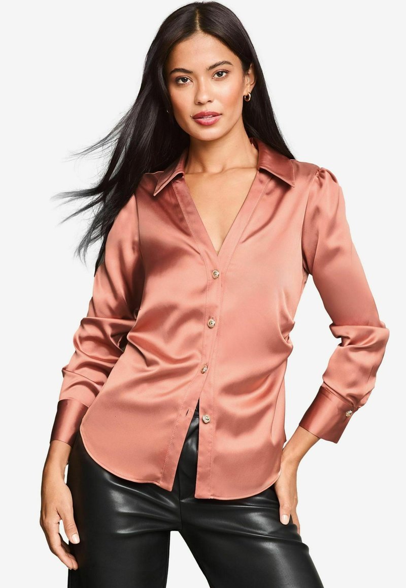 Blusa di raso rosa con scollo a V, maniche lunghe, bottoni visibili e orlo curvo; abbinata a pantaloni in pelle nera.